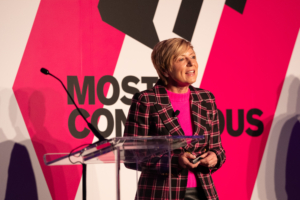 Susan Howe on stage at Most Contagious New York. She is in a plaid jacket and bright top standing in front of a large backdrop with the Most Contagious logo on it.